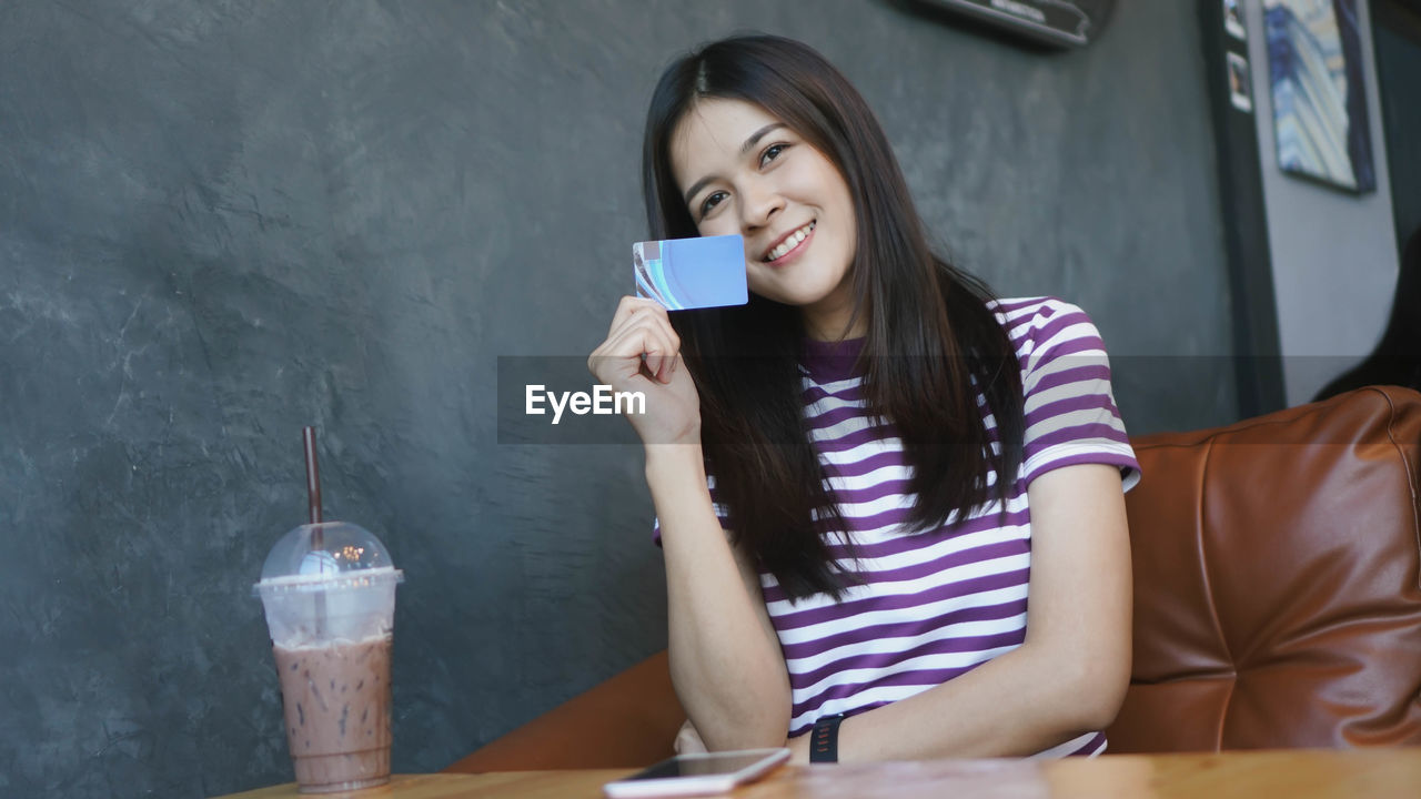 Portrait of smiling young woman holding credit card sitting at table with coffee in cafe