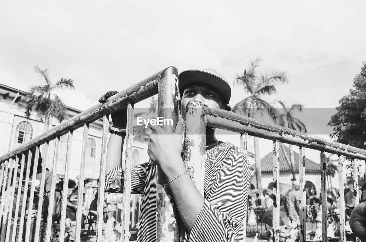 Young man standing by gate against sky