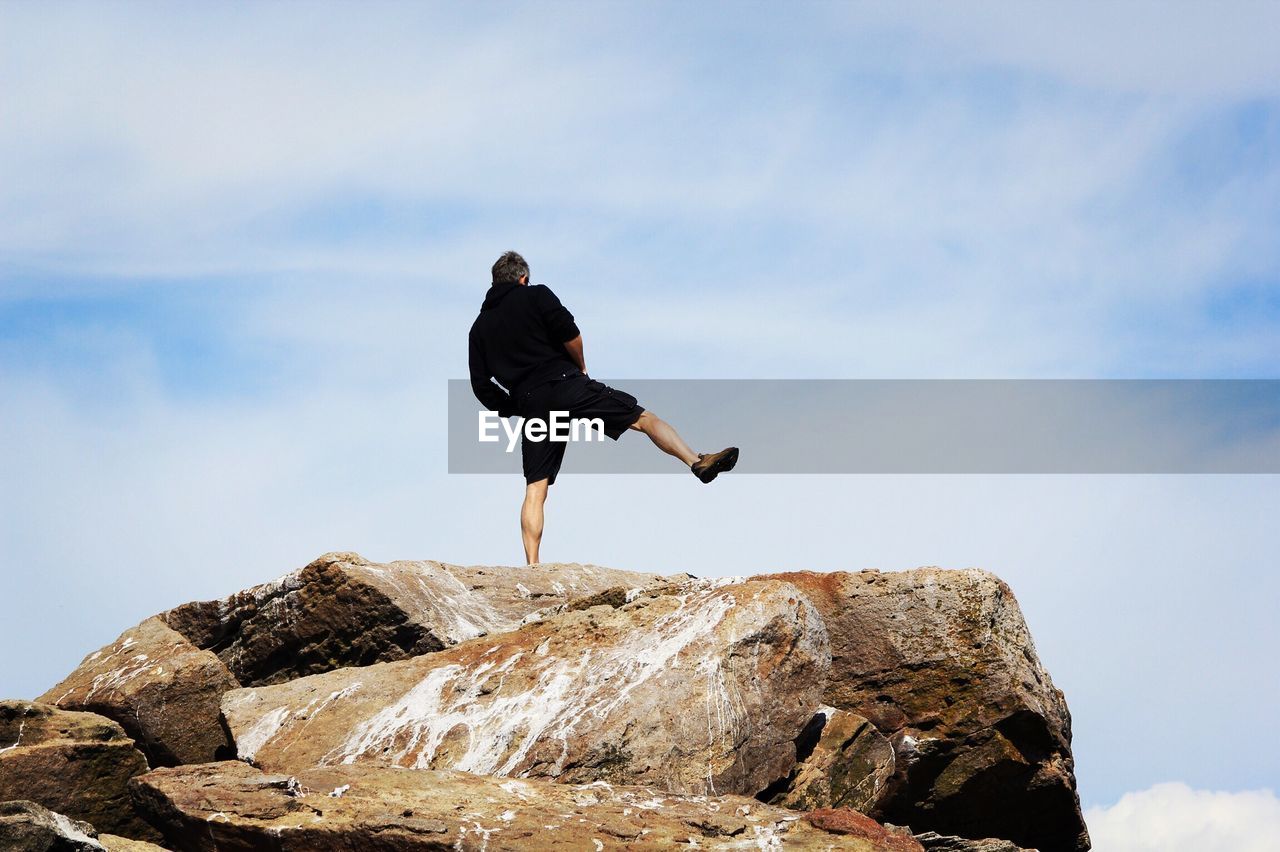 Rear view of man standing on one leg at rock against sky