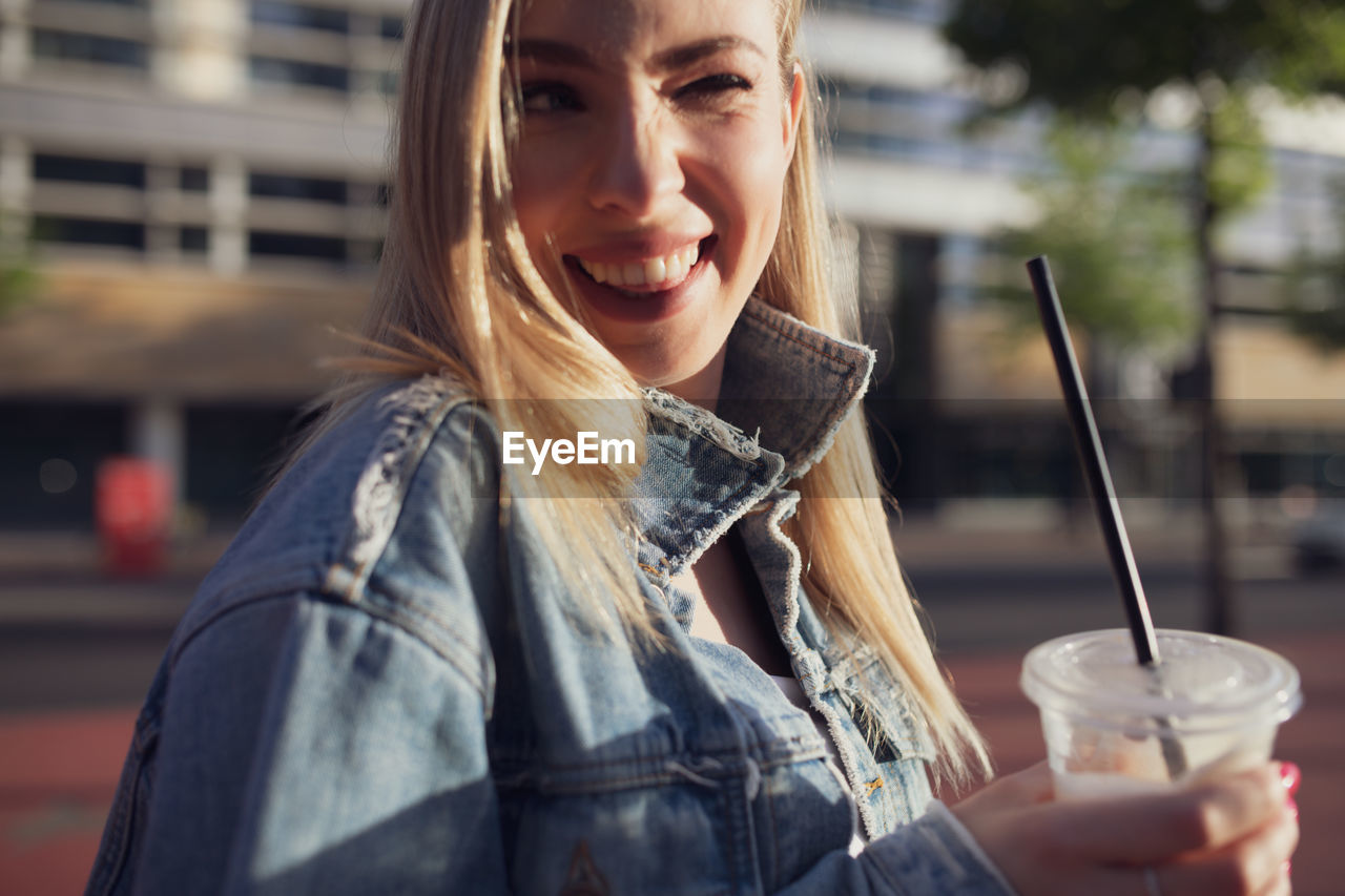 Young woman winking while holding disposable cup in city