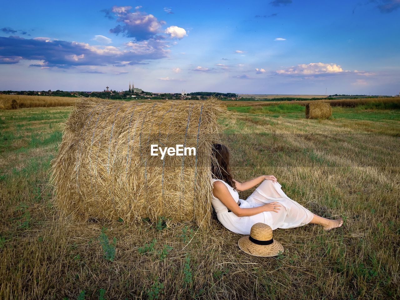 Rural scene of woman sitting down on the ground and leaning against a haystack on the field