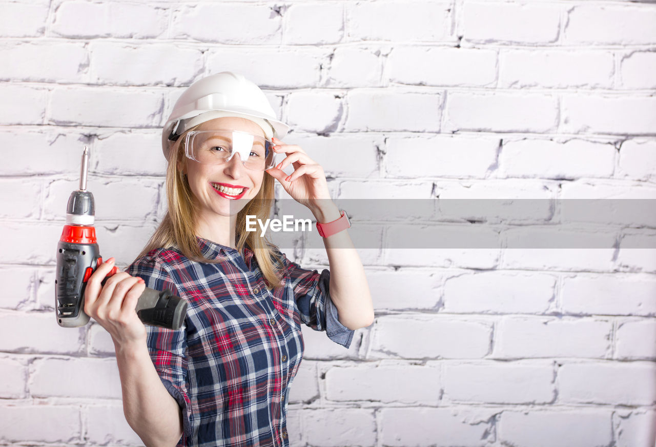 Portrait of smiling young woman holding drill while standing against wall