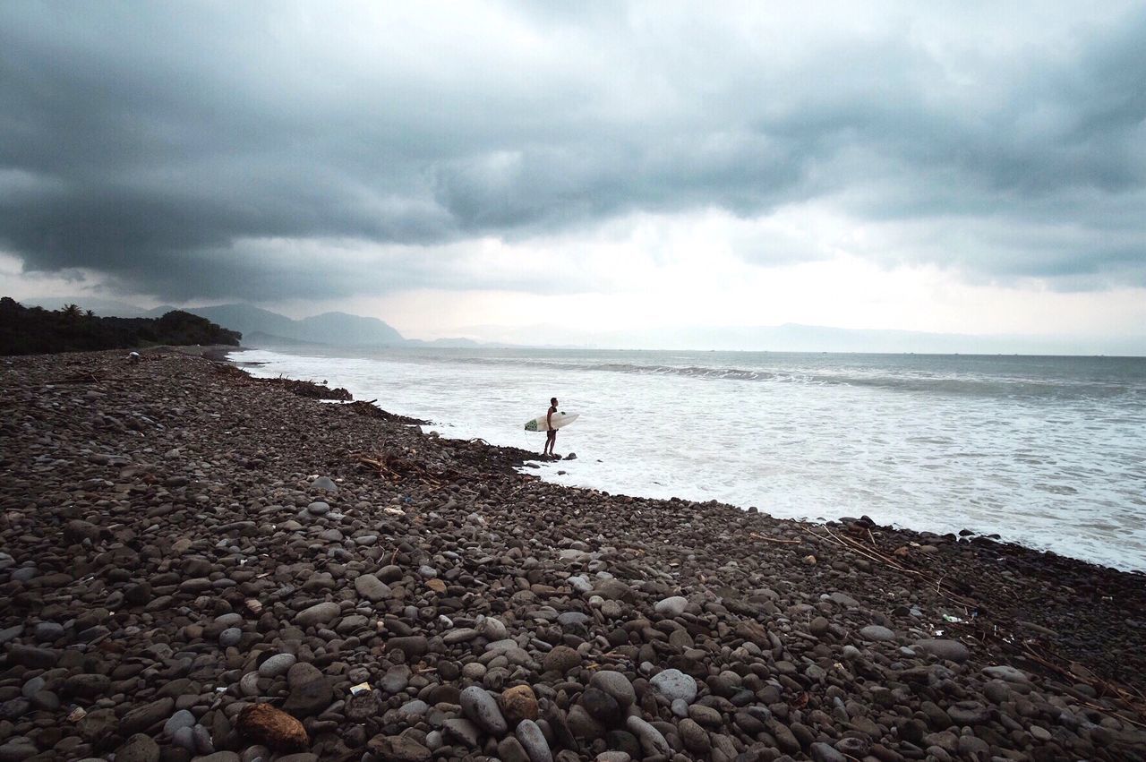 Man standing with surfboard at shore against cloudy sky