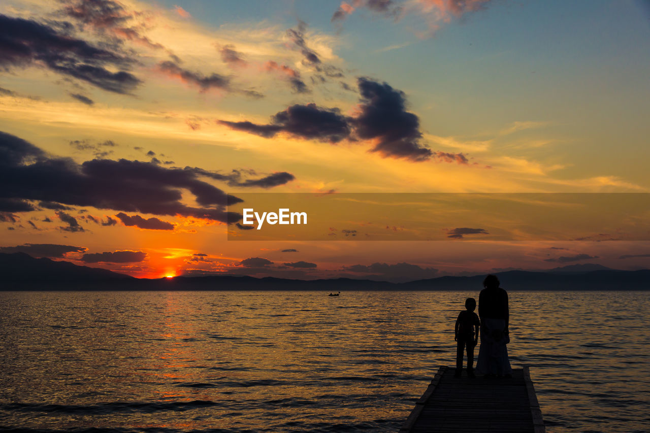 SILHOUETTE PEOPLE STANDING ON SHORE AGAINST ORANGE SKY