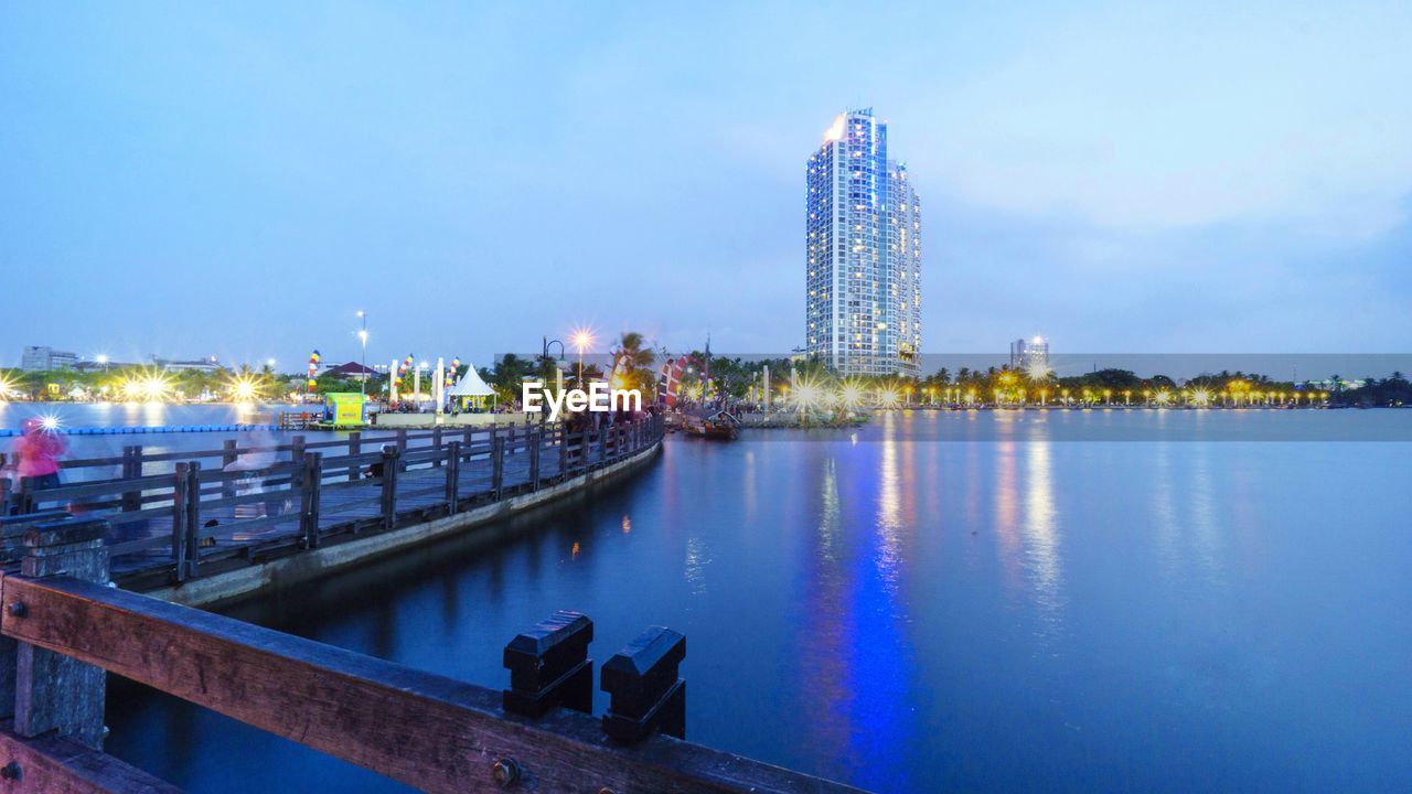 ILLUMINATED BUILDINGS BY RIVER AGAINST SKY AT DUSK