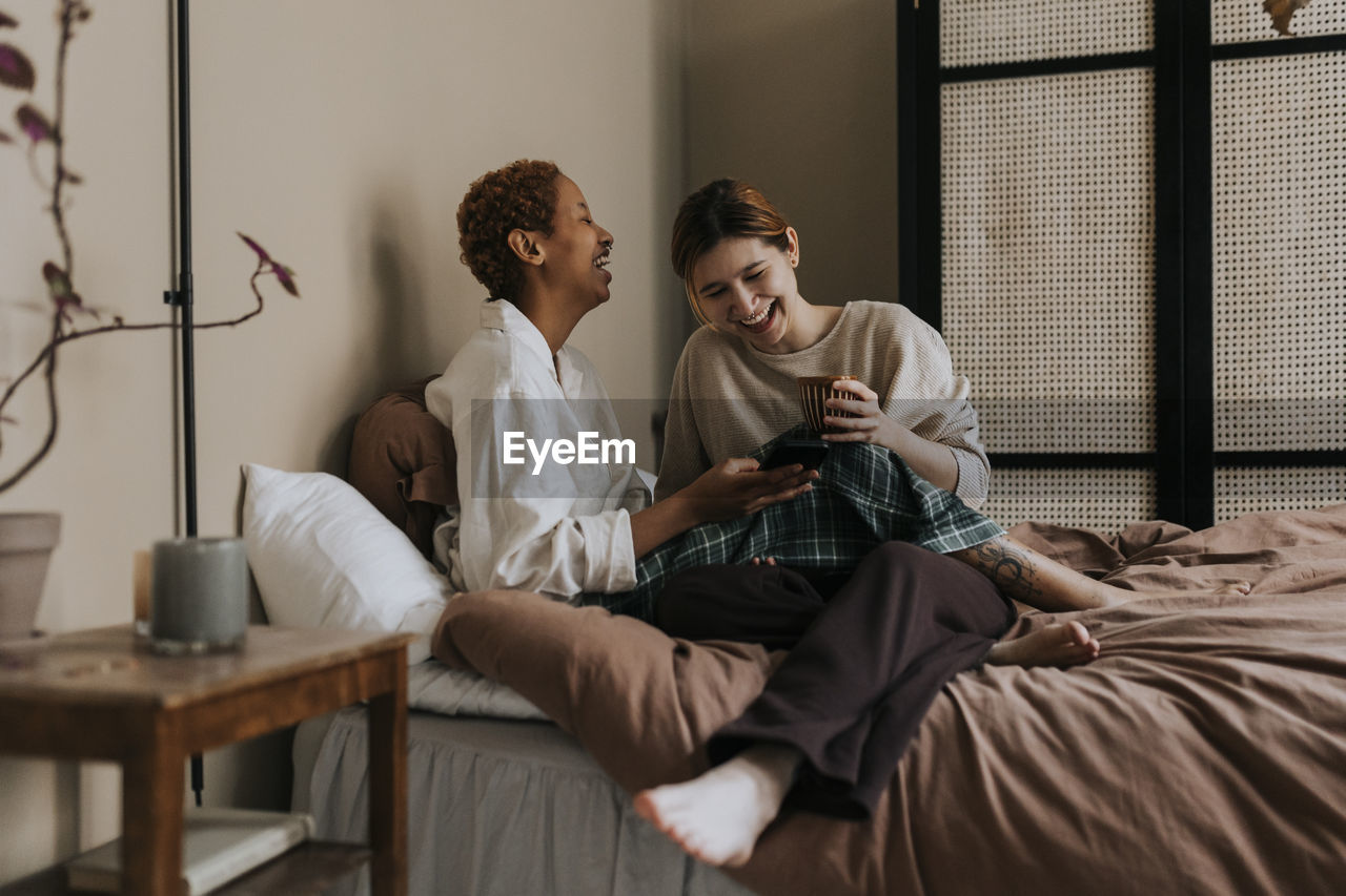 Happy non-binary couple sharing smart phone while sitting on bed at home