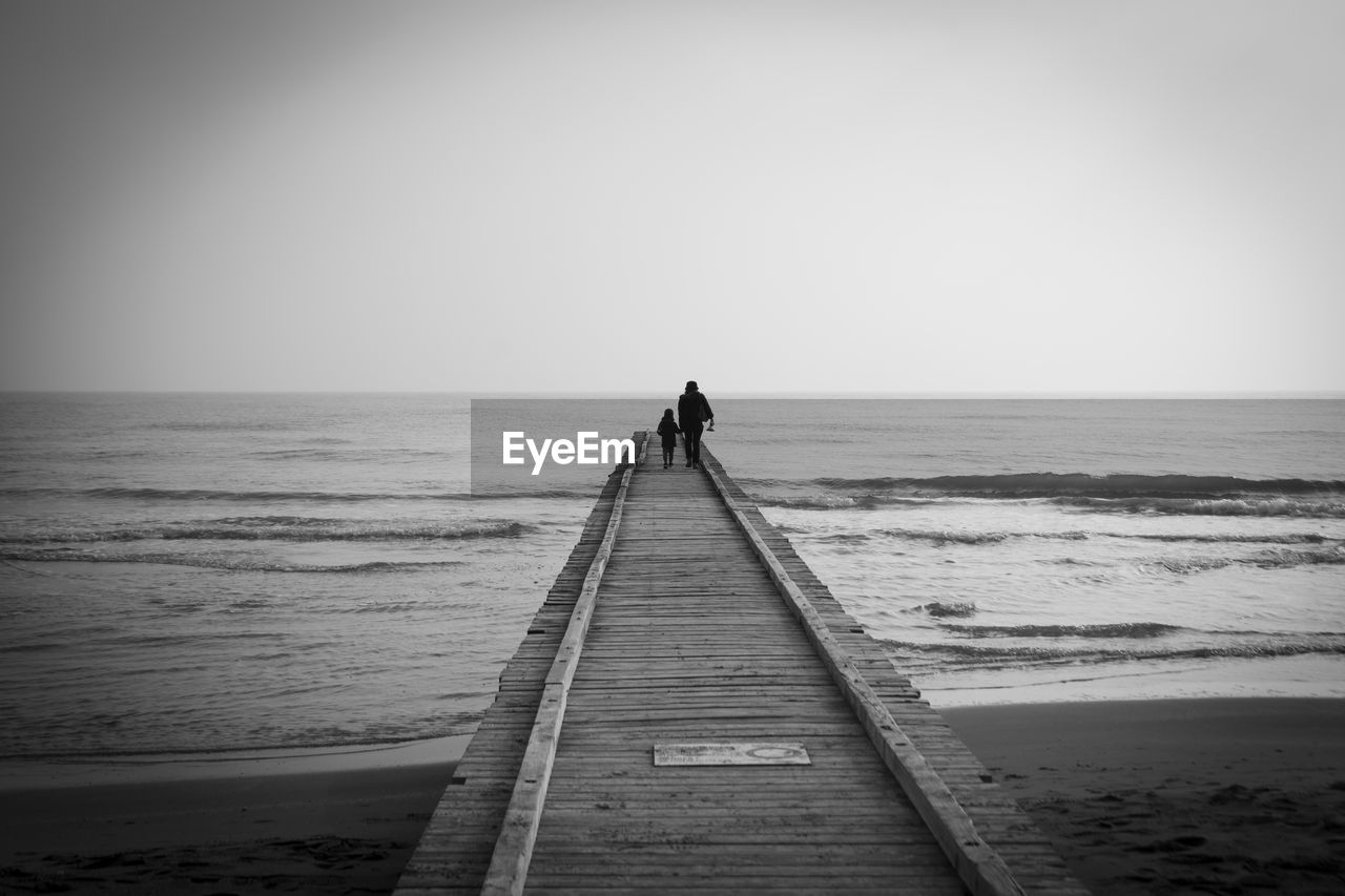 sea, water, horizon over water, horizon, sky, beach, land, black and white, nature, white, monochrome, monochrome photography, scenics - nature, beauty in nature, tranquility, ocean, tranquil scene, one person, wave, the way forward, holiday, black, clear sky, rear view, vacation, pier, trip, shore, leisure activity, full length, coast, lifestyles, copy space, adult, solitude, outdoors, wood, idyllic, day, sand, morning, men, non-urban scene, motion, travel, silhouette, travel destinations, relaxation, urban skyline, body of water, architecture, jetty, walking, women, diminishing perspective