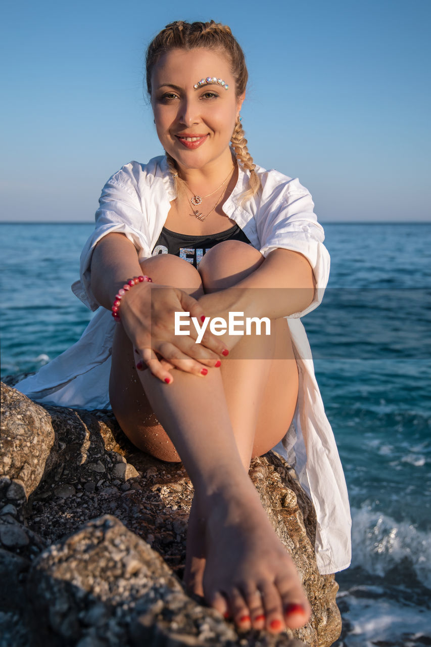 YOUNG WOMAN SITTING ON ROCK AGAINST SEA