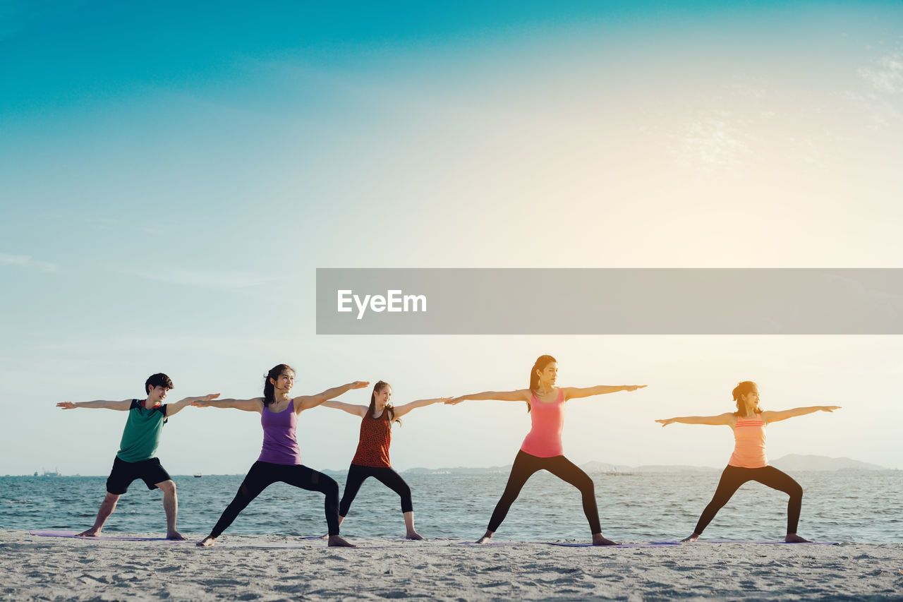 Young friends practicing yoga in warrior position on shore at beach during sunny day