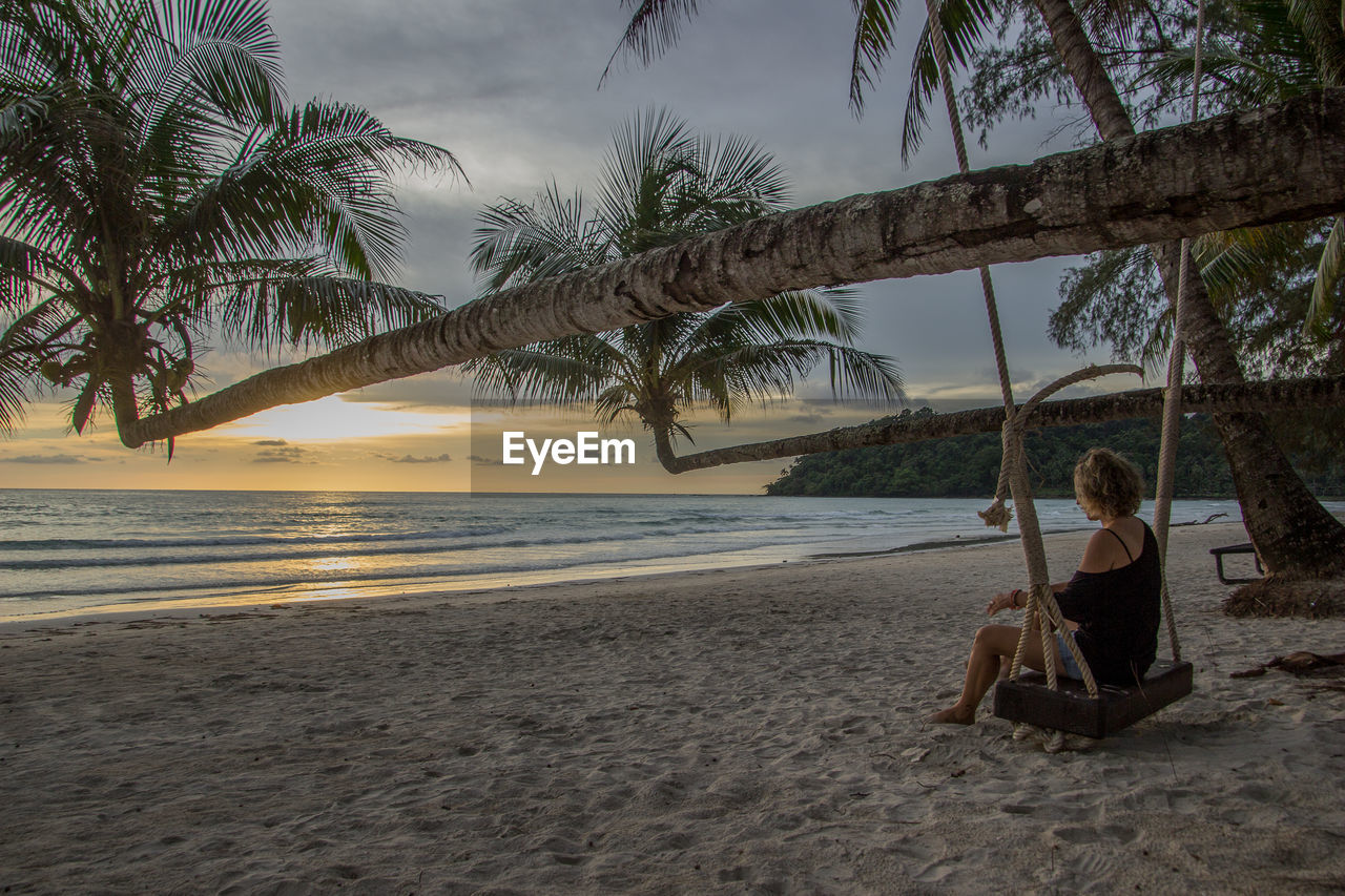 Woman sitting on swing at beach against sky during sunset