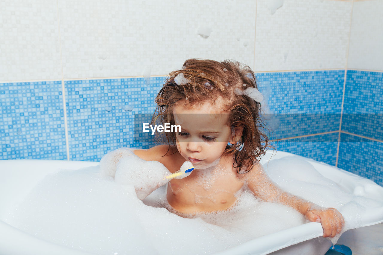 Topless girl brushing teeth while sitting in bathtub at home