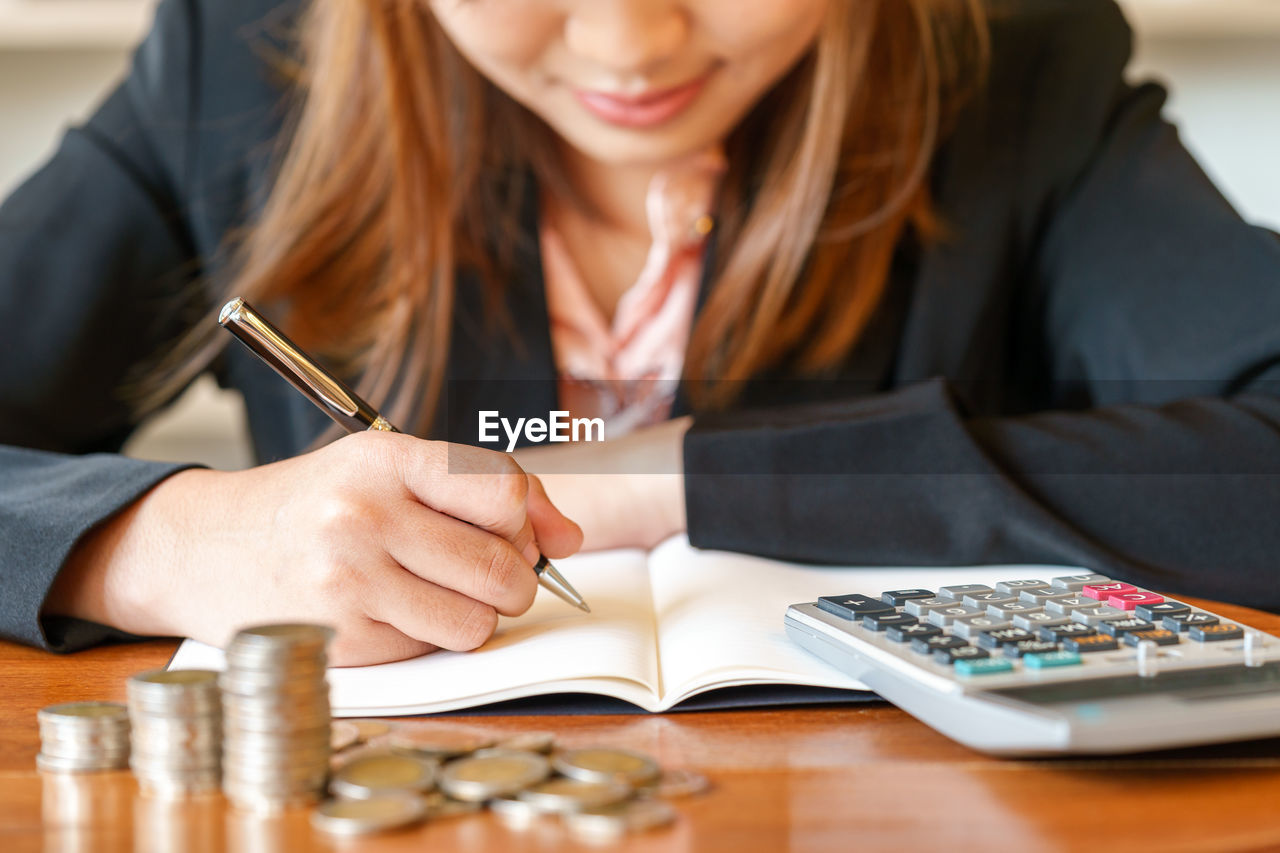 Midsection of young woman writing in diary by coins and calculator on desk