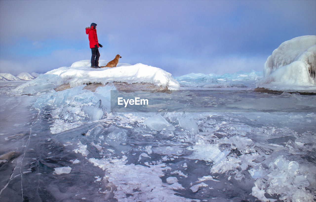 Person with dog on snow covered rock during winter against cloudy sky