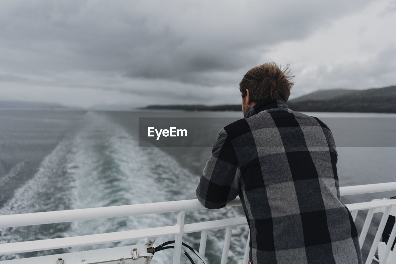 Rear view of man leaning on railing of boat in sea against sky