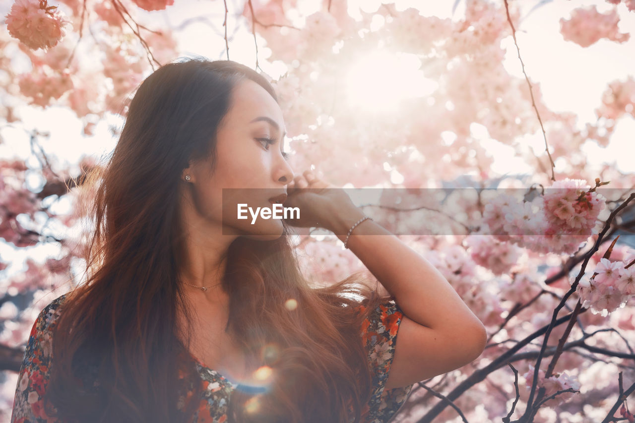 Young asian woman, in a floral pattern dress, in front of blossoming cherry tree in the spring time