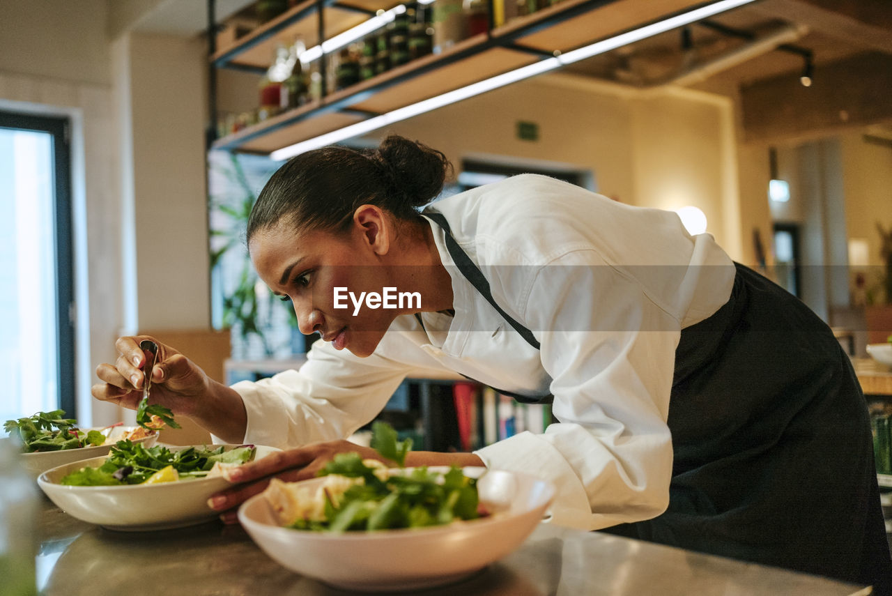 Focused female chef arranging cilantro on salad bowl using tweezers in commercial kitchen