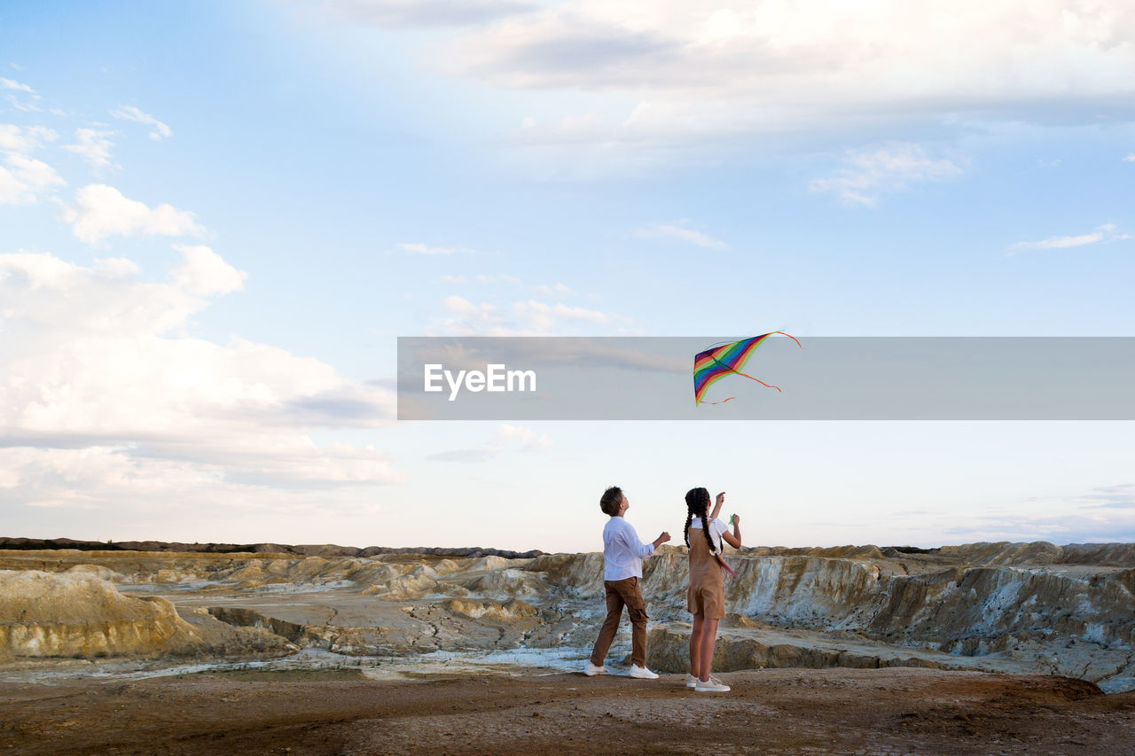 A boy and a girl launch a bright kite into the sky in the mountains near the river.