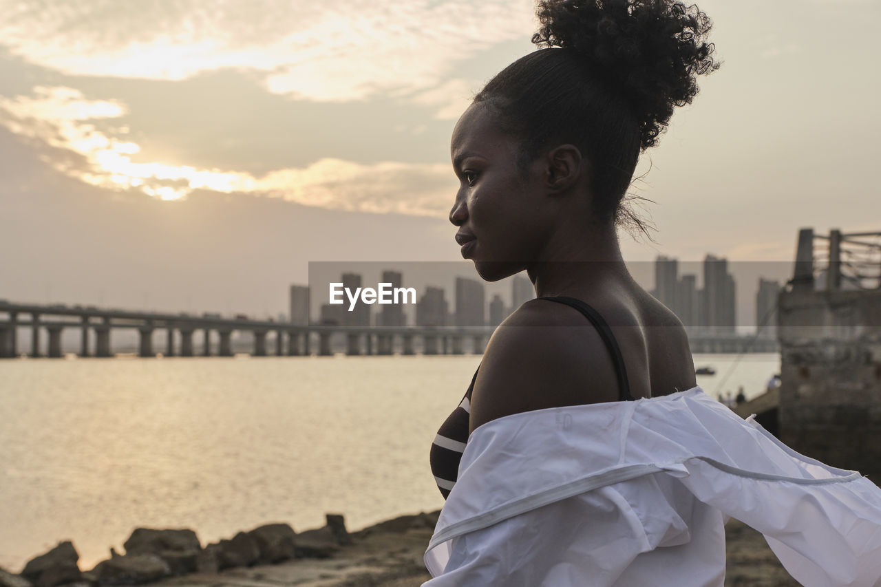 Tranquil pensive young african american female in casual clothes relaxing on urban seafront at sunset time