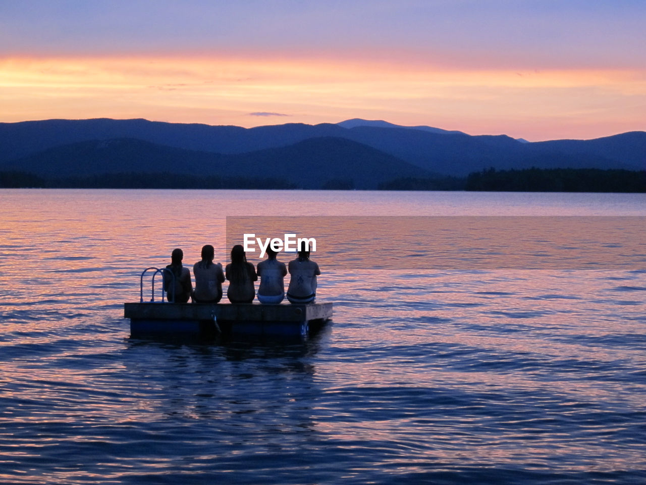 Rear view of women sitting on wooden raft in sea against sky during sunset