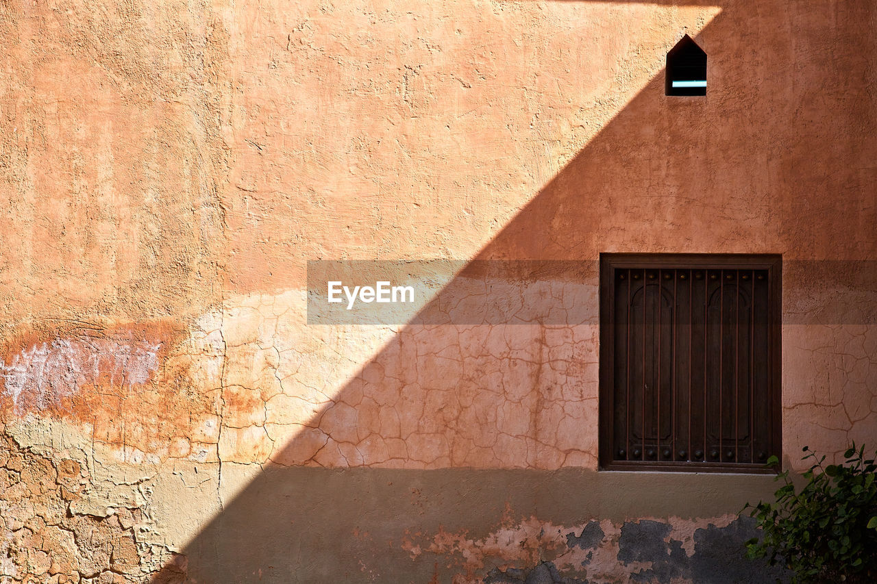 A weathered exterior house wall, with a closed window and a shadow cutting it in two