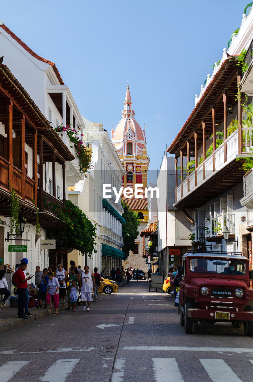 architecture, town, building exterior, city, built structure, street, transportation, road, neighbourhood, mode of transportation, building, travel destinations, car, motor vehicle, city street, sky, travel, tourism, nature, city life, vacation, downtown, urban area, clear sky, infrastructure, day, group of people, vehicle, sunny, cityscape, town square, crowd, outdoors, residential district, history, religion, the past, lane, place of worship, residential area, adult, road marking, sunlight, walking