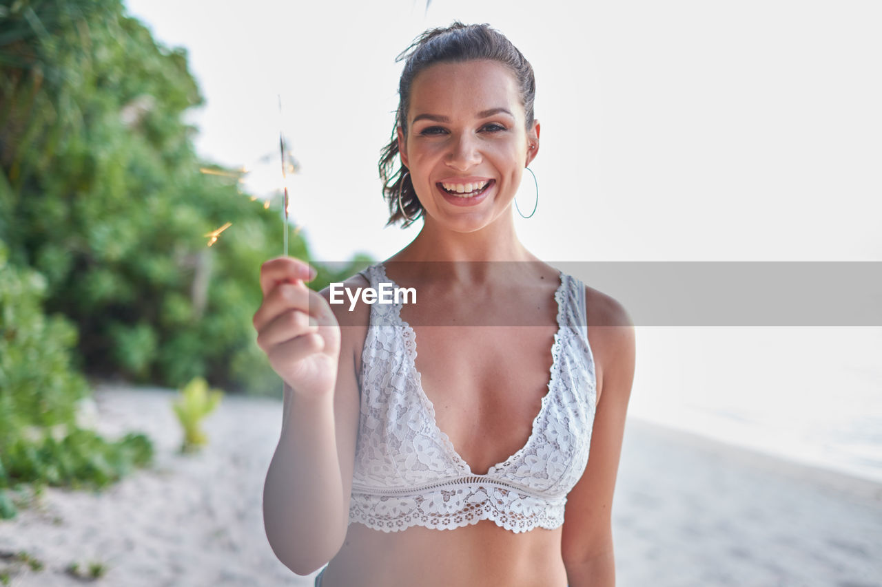 Portrait of smiling young woman holding illuminated sparkler while standing at beach