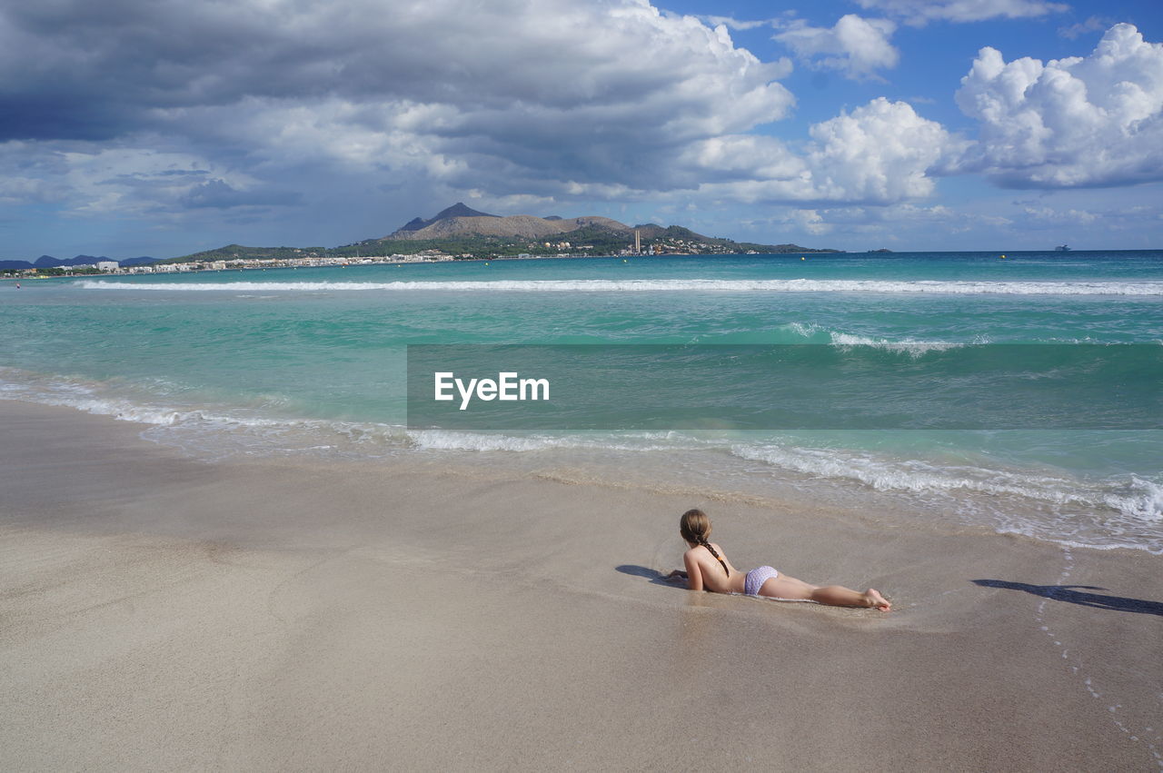 Full length of girl lying on shore at beach against cloudy sky