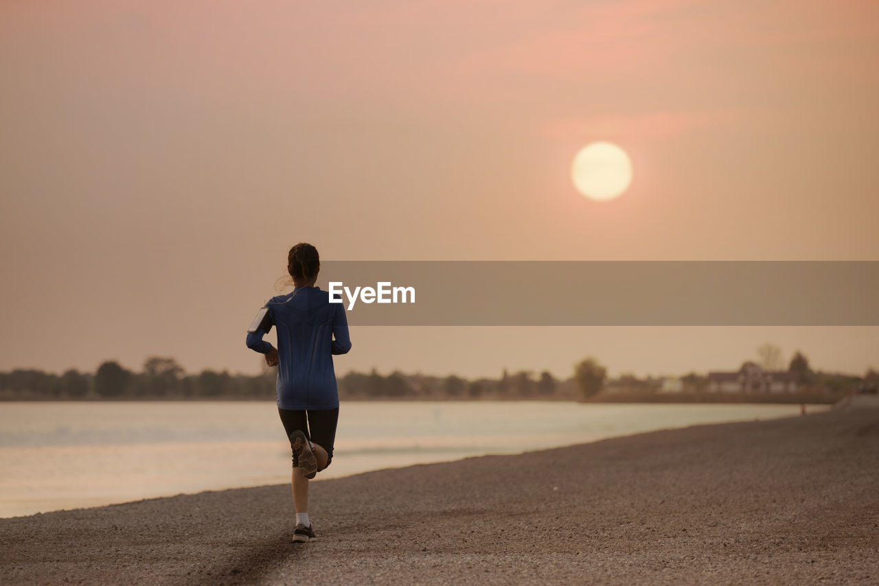 Rear view of woman running at beach against sky during sunset