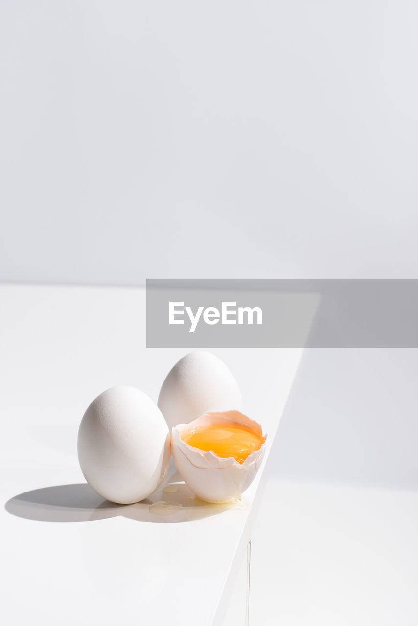 High angle of whole and broken egg with yolk in shell placed on edge of table on white background in studio