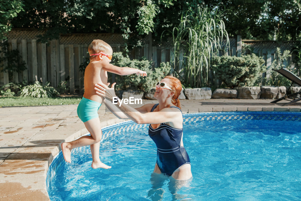 Caucasian mother training toddler son to swim in swimming pool outdoor. baby boy diving 