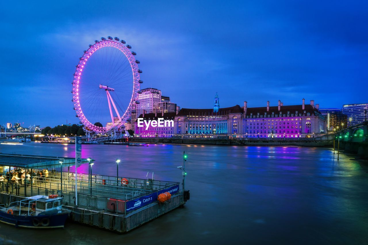 Illuminated ferris wheel by river against sky in city