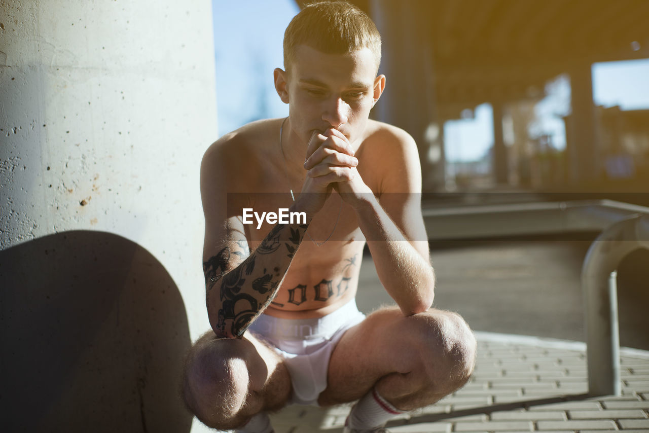 Thoughtful shirtless young man crouching outdoors