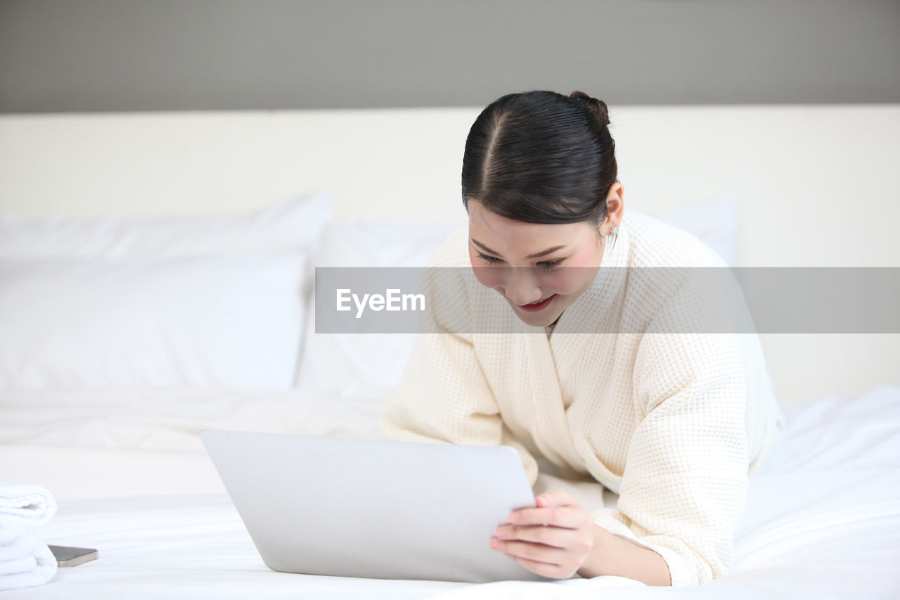 YOUNG WOMAN USING MOBILE PHONE WHILE SITTING ON BED