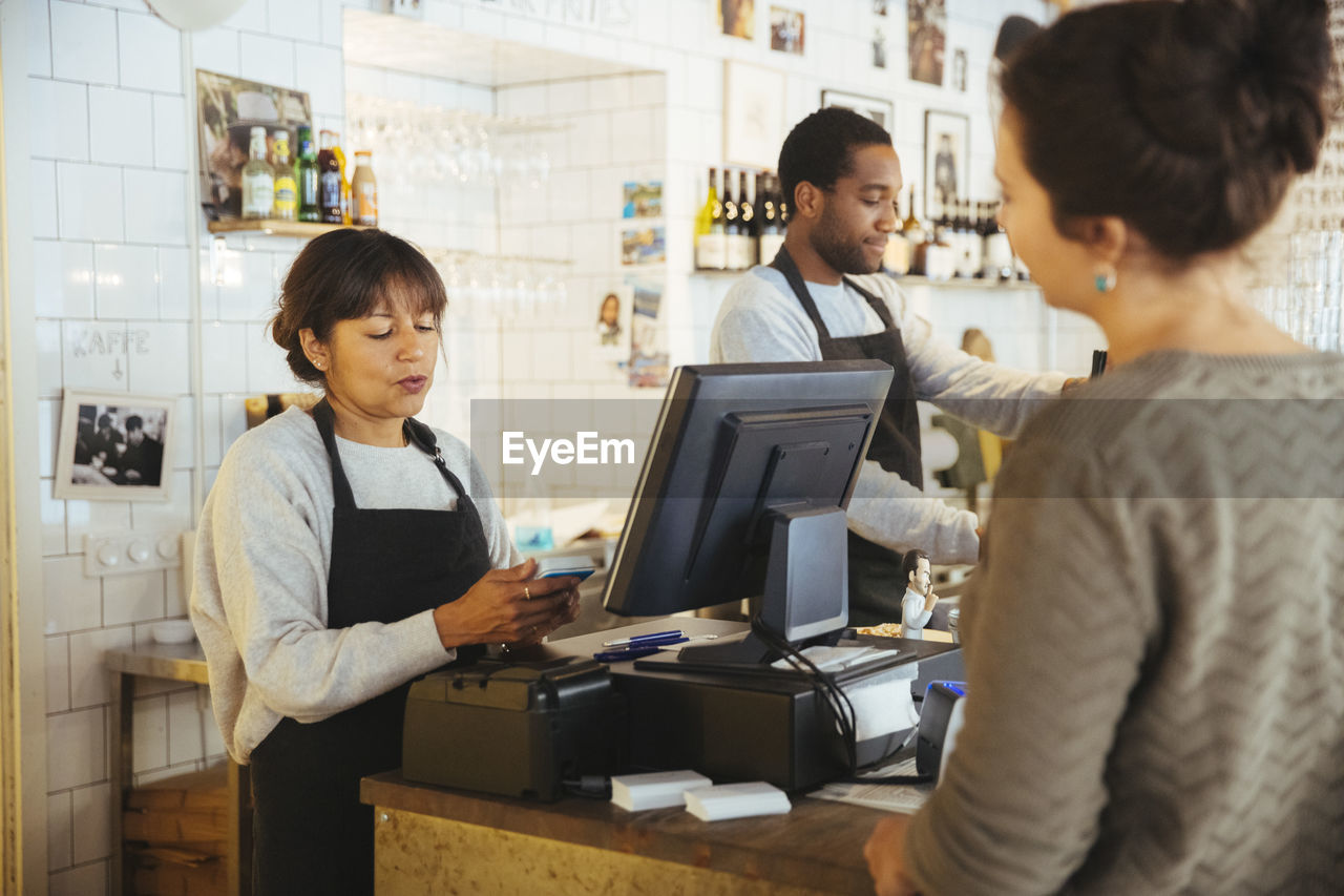 Mature female employee billing customer standing at checkout counter in delicatessen