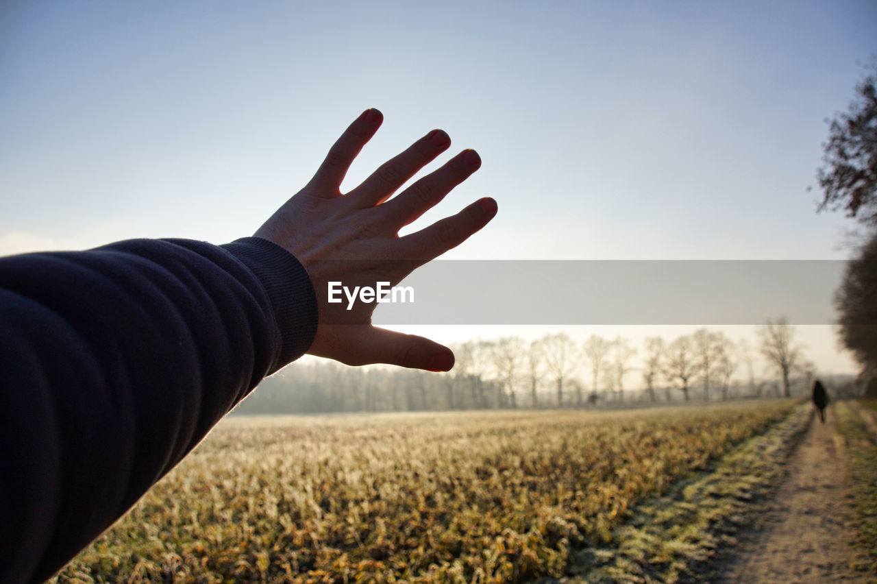 Cropped hand of man blocking sunbeam on field against sky