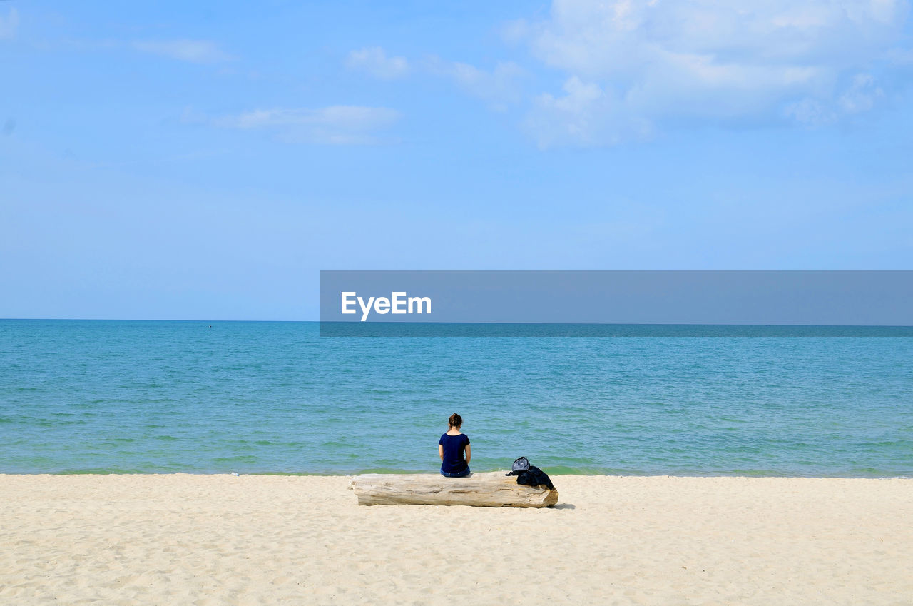 Rear view of woman sitting on wood at sandy beach against blue sky