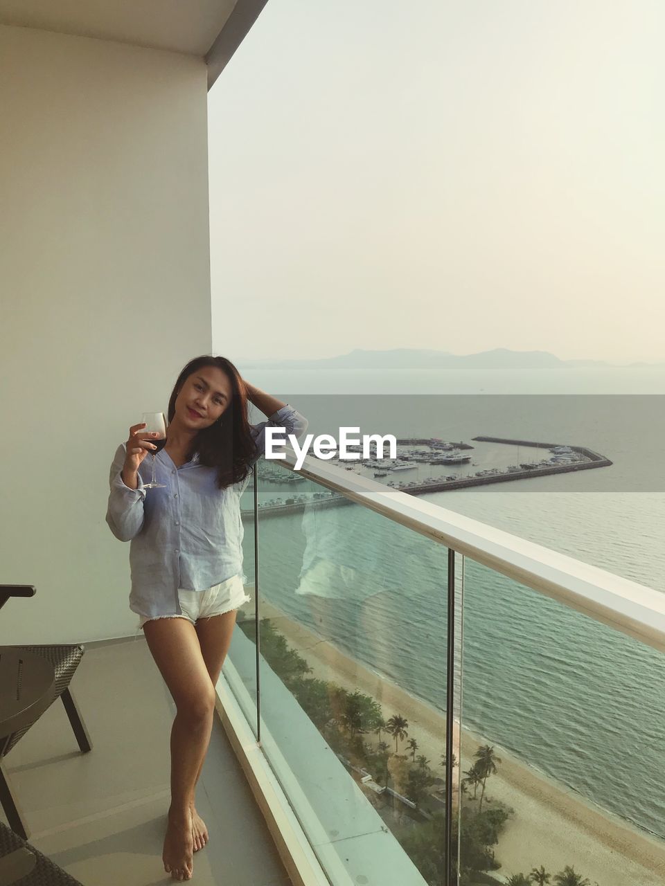 Portrait of smiling woman with wineglass standing at balcony by sea