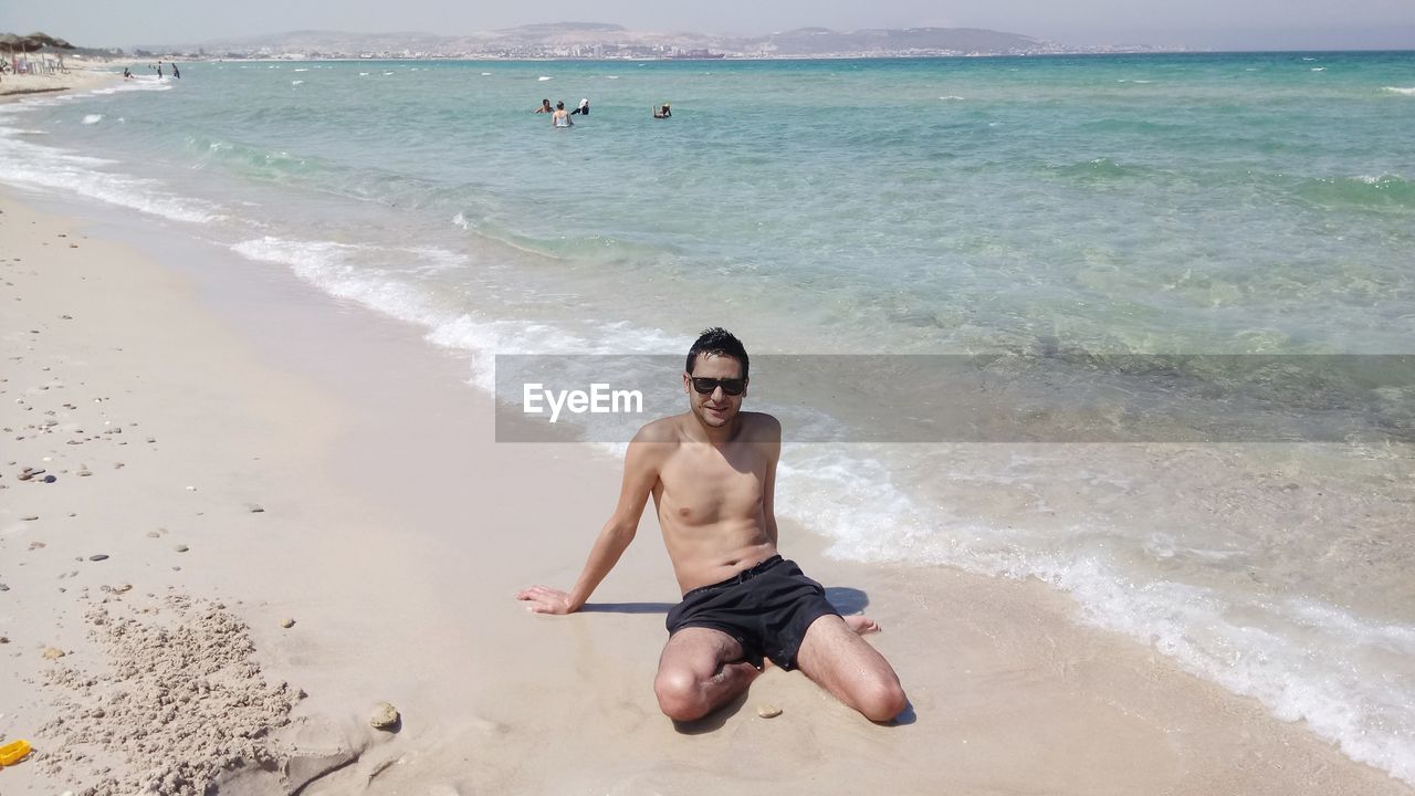 Full length of shirtless man sitting on shore at beach
