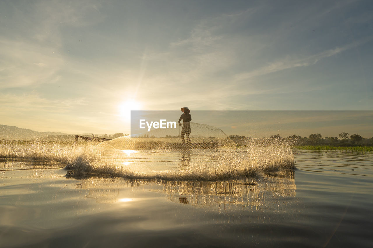 MAN STANDING ON LAKE AGAINST SKY DURING SUNSET