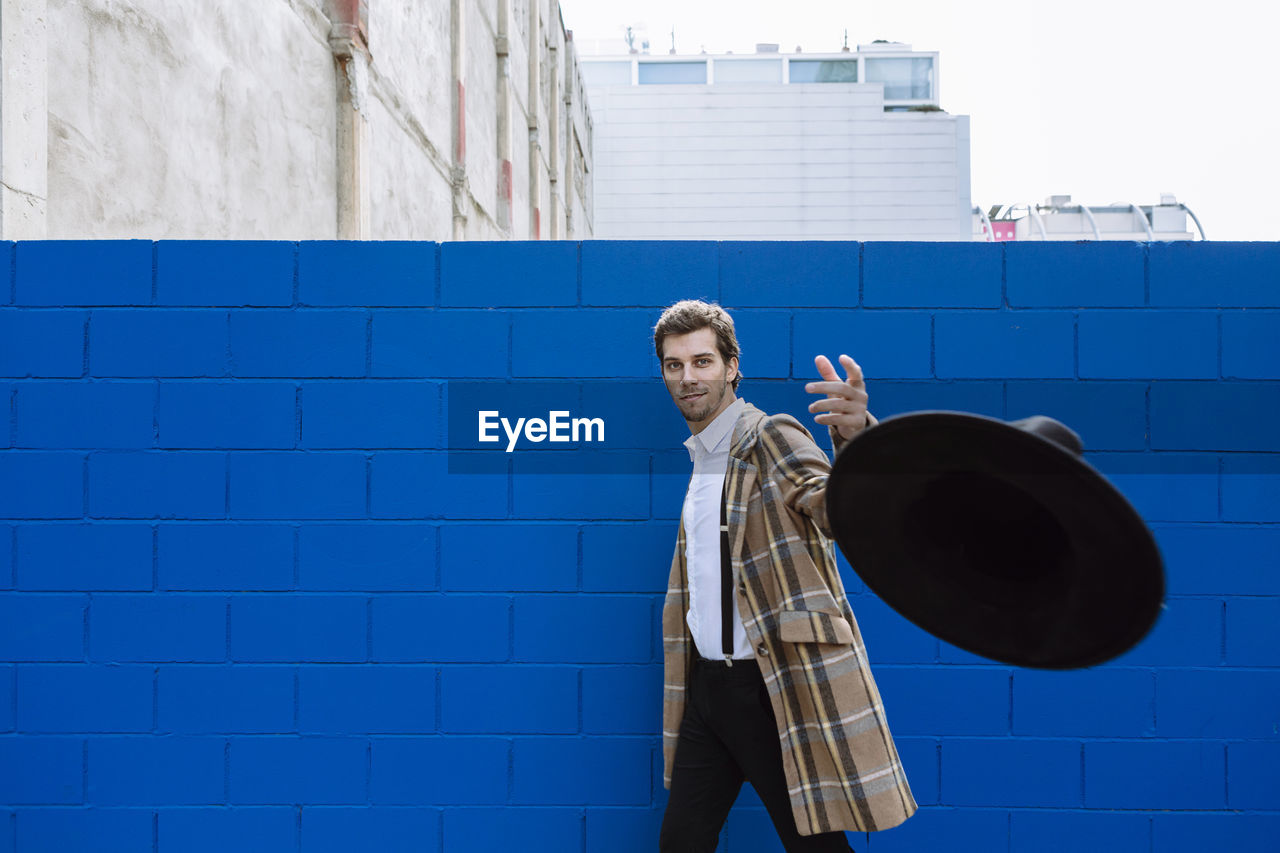 Stylish man wearing checked jacket throwing black hat against blue wall