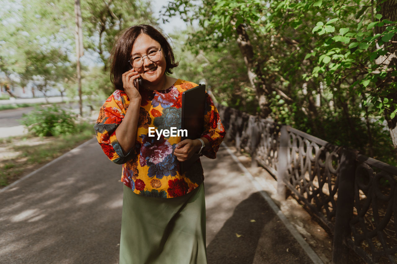 Cheerful young woman with glasses talking on a mobile phone in lush green park.