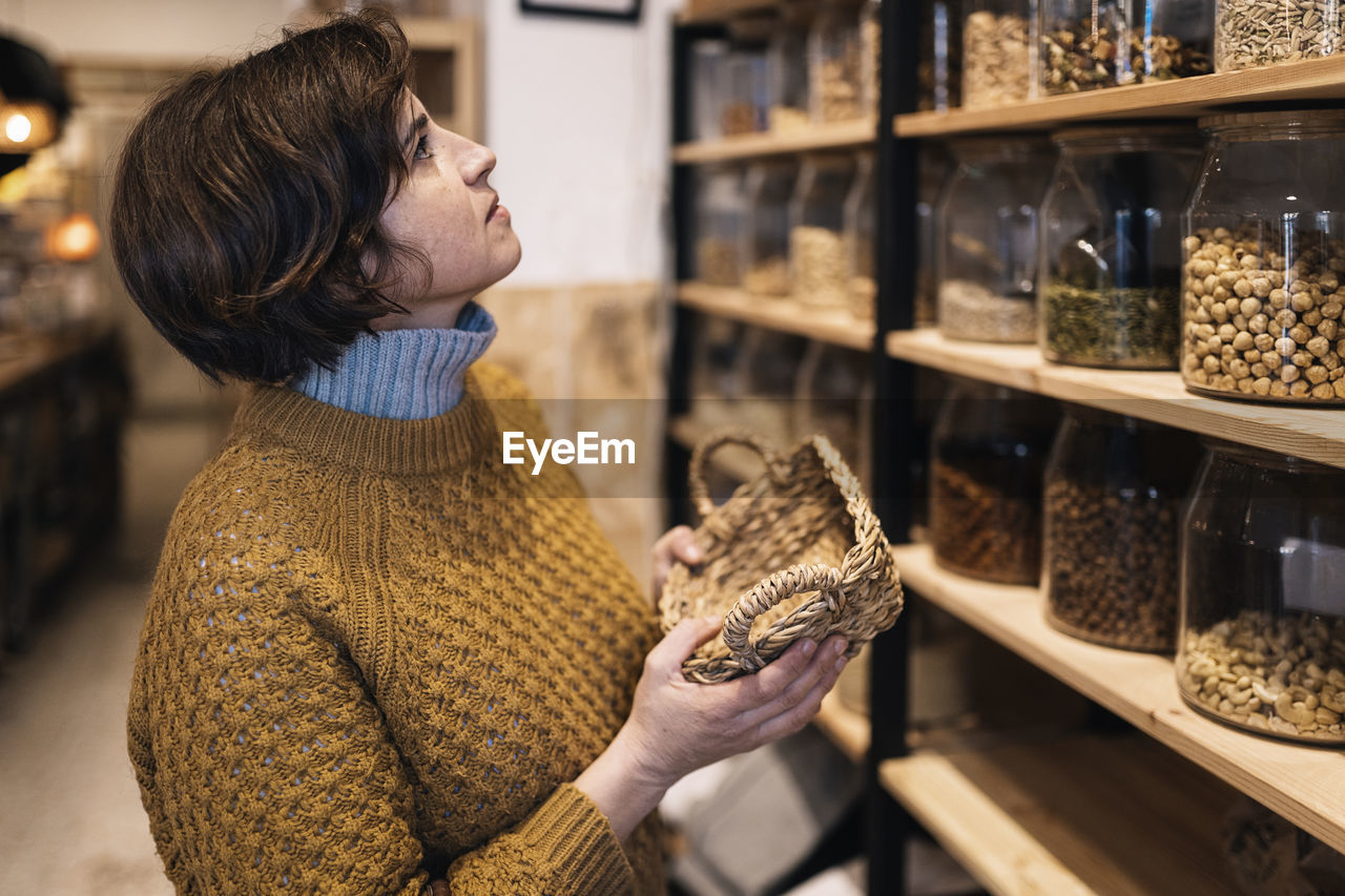 Side view of calm female with short brown hair in yellow knitted sweater standing with wicker basket in hands near shelves with jars with nuts in eco friendly shop