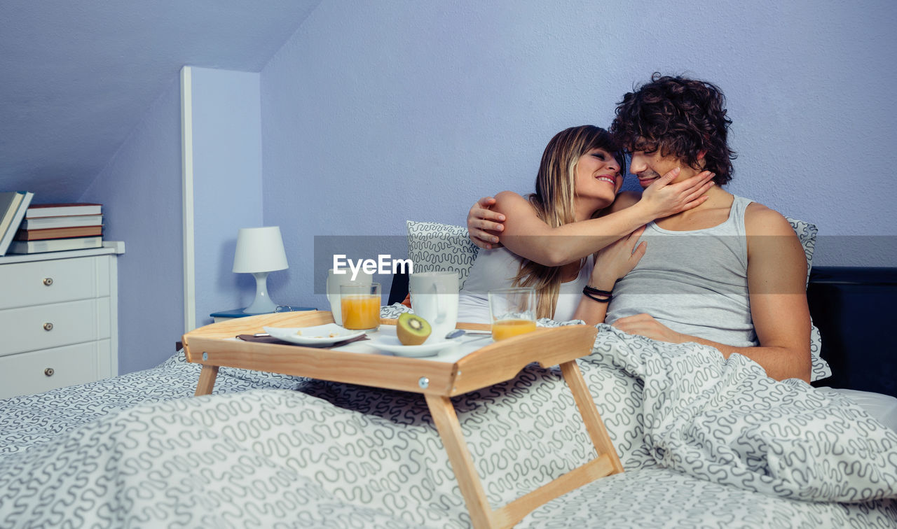 Young couple with food and drinks on bed at home