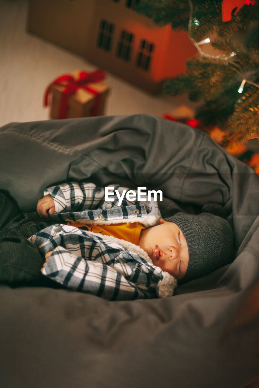 A cute newborn baby in a plaid jacket and a knitted hat is sleeping on a pouf next to a christmas 