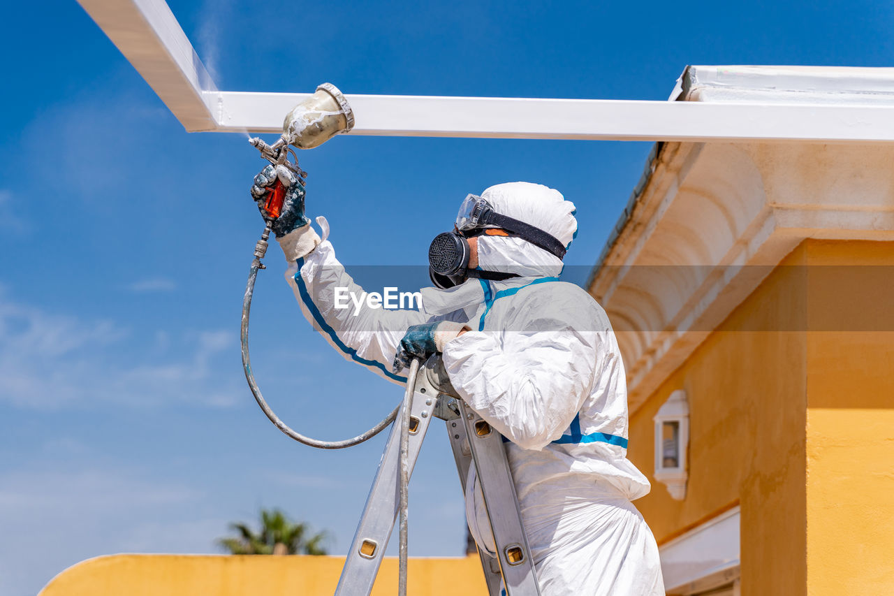 Anonymous employee in protective uniform and mask standing on ladder and using air brush to paint frame structure against cloudy sky on sunny day
