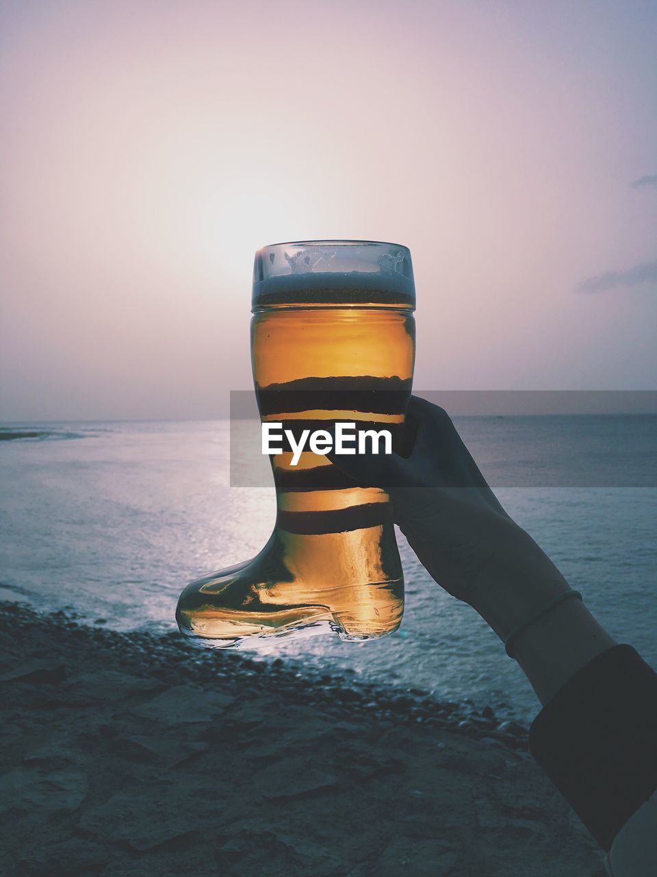 Cropped hand of woman holding boot shape beer glass at beach against sky during sunset
