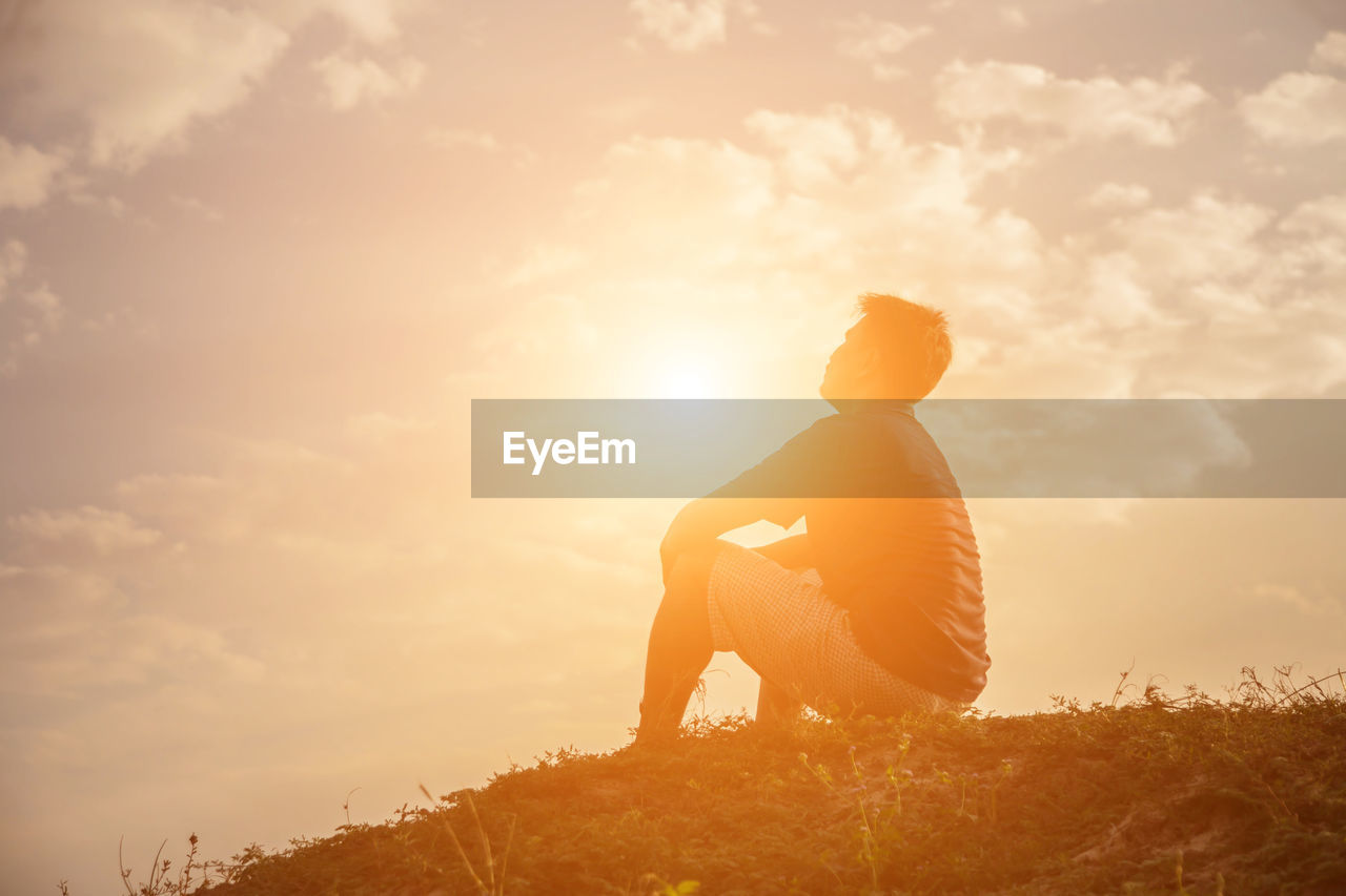 Side view of man sitting on land against sky during sunset