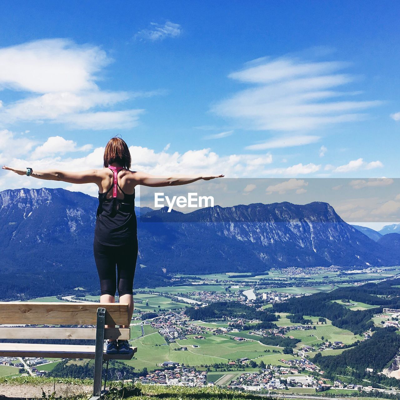 Rear view of woman standing on bench with arms outstretched by mountains against sky