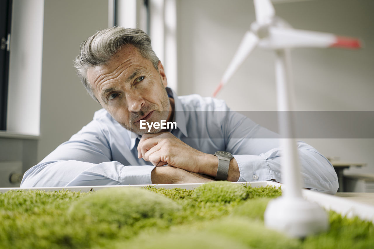 Businessman looking at wind turbine toy over moss frame while leaning on table at office
