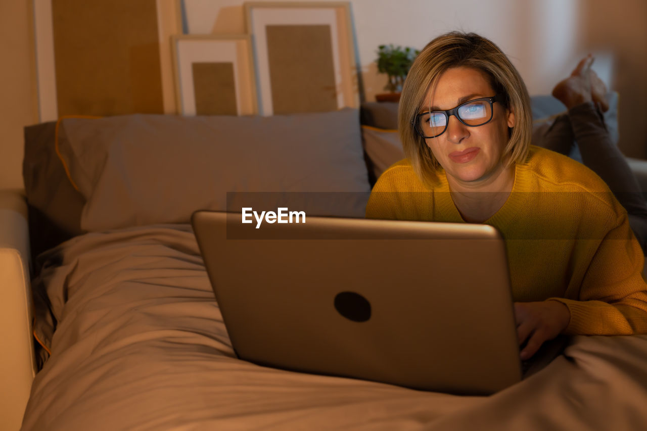 portrait of young woman using laptop while sitting on bed at home